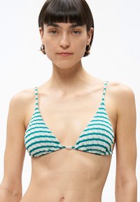 Νέα γυναίκα με κοντά σκούρα μαλλιά, φοράει χρυσούς κρίκους στα αυτιά και πράσινο-άσπρο ριγέ υφασμάτινο μαγιό με υφή, σε λευκό φόντο.