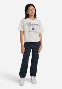 Gaiši pelēka īsām piedurknēm T-krekls ar tumši zilām burtiem; savienots ar tumši zilām kravas biksēm un baltām kedām. Vienkāršs, ikdienišķs apģērbs.