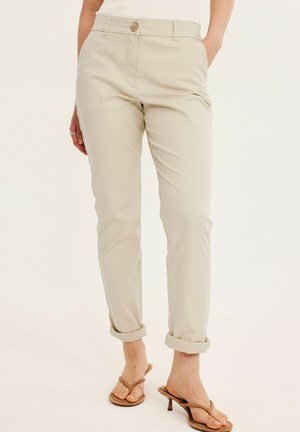 Kvinde iført beige opbukte chinos og beige hælede sandaler, stående med hænderne i lommerne mod en ensfarvet baggrund.