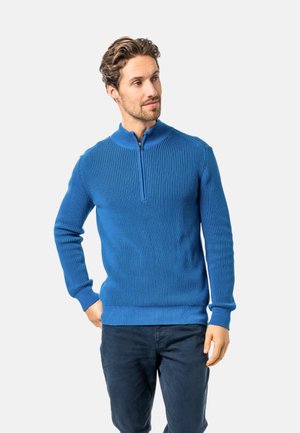 Mann trägt einen blauen Strickpullover mit Reißverschluss und dunkle Hosen, steht mit einer Hand in der Tasche und schaut seitlich vor einem einfarbigen Hintergrund.