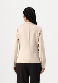 Blazer beige aderente con design strutturato, maniche lunghe e una cucitura posteriore singola, abbinato a pantaloni neri a gamba larga.