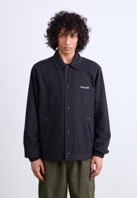 Black jacket with a collar, featuring front buttons, two side pockets, elastic cuffs, and a small embroidered logo on the chest.