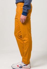 Oranje katoenen broek met een slanke pasvorm, voorzien van zijzakken en een knoopsluiting. Gecombineerd met witte sneakers met rode en blauwe accenten.