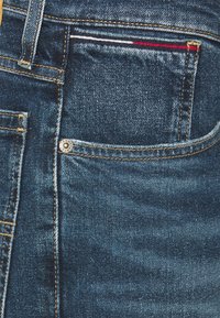 Jeans de mezclilla azul oscuro con una textura ligeramente desgastada. Presentan un bolsillo frontal con costuras decorativas y un remache metálico para mayor durabilidad.