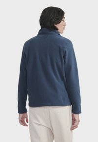 Marineblå fleece-pullover med høj krave og lange ærmer. Blød struktur og enkelt design. Har et diskret logo på ryggen.