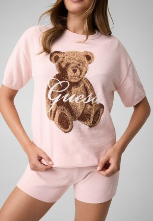 Mujer con camiseta de manga corta rosa claro y pantalones cortos a juego con un oso de peluche y el logo "Guess" en la parte delantera de la camiseta.