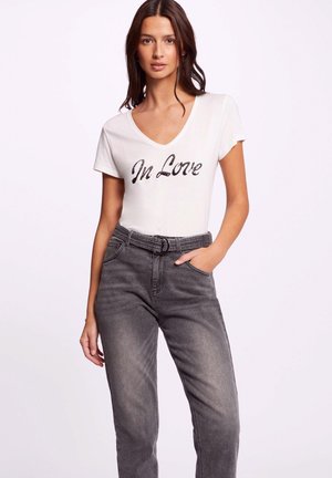 Witte v-hals t-shirt met zwarte pailletten tekst "In Love." Gecombineerd met hooggeknoopte, vergrijsde spijkerbroek met een riem.