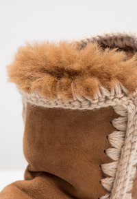 Botte en daim marron avec une doublure en fourrure beige duveteuse. Présente des accents tricotés beiges le long du bord, mettant en valeur sa texture et son savoir-faire.