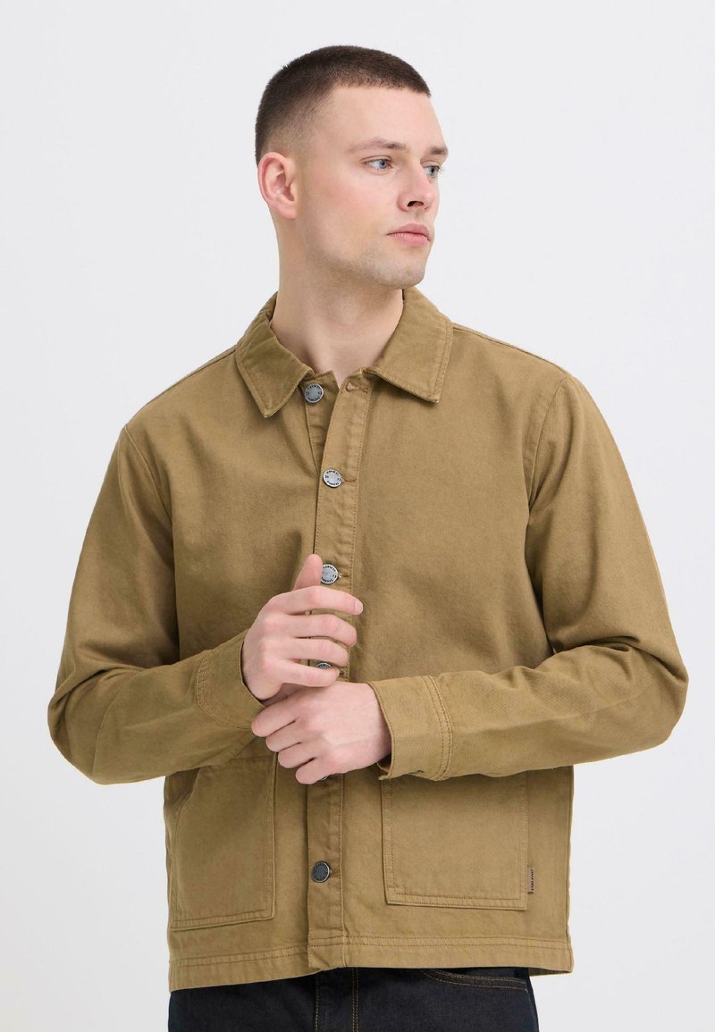 Νέος άνδρας με ανοιχτό μπεζ σακάκι που έχει κουμπώσει, κοιτάζει προς τα πλάγια με τα χέρια κοντά στο στήθος σε λευκό απλό φόντο.