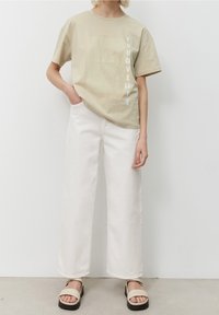 Beige bomullströja med grafisk design, vit text som läser "YOU GIVELIFE," i kombination med vida vita byxor och ljusa sandaler.