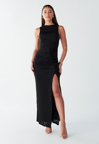 Čierne dlhé šaty bez rukávov so získaným nariasením, bočným rozparkom a jemnou textúrou. Modelka má na sebe sandále, ktoré zdôrazňujú elegantný strih šiat.