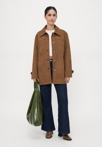 Braune Wildlederjacke mit spitzem Kragen, sechs vorderen Knöpfen und langen Ärmeln; kombiniert mit dunkelblauen weiten Jeans und einer grünen Tasche.