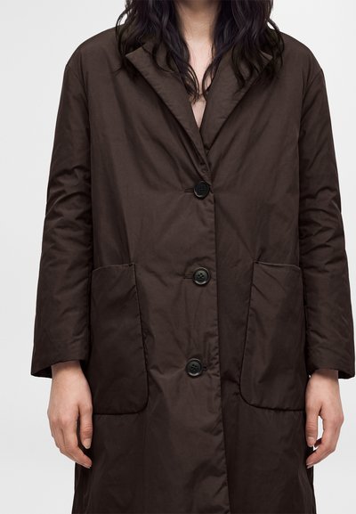 Personne portant un long manteau brun foncé avec trois boutons noirs et de grandes poches avant, visible du cou jusqu'au milieu de la cuisse.