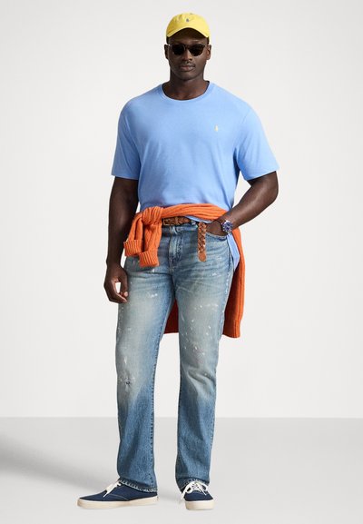 Šviesiai mėlynas marškinėlis, laisvo kirpimo džinsai su nudrengtais elementais, oranžinis megztinis, pririštas juosmenyje, geltona kepurė, juodi akiniai nuo saulės ir mėlyni batai.