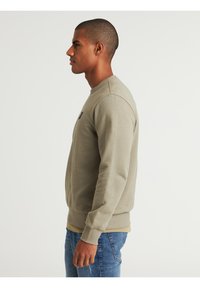 Sudadera verde oliva con cuello redondo y puños de canalé. Presenta un pequeño logo en el pecho. Combinada con jeans azules. Textura suave.