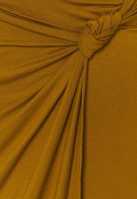 Tissu jaune moutarde à la texture douce, comportant des plis drapés et un détail noué, créant un intérêt visuel dynamique et de la profondeur.