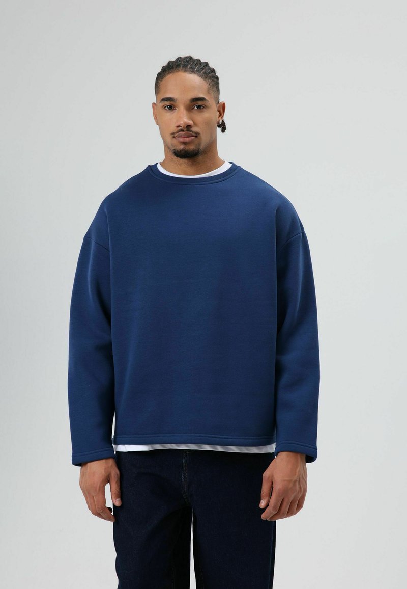Marineblauwe sweatshirt met een ronde hals, aflopende schouders en geribde manchetten, gedragen over een wit shirt, gecombineerd met donkere denim jeans.