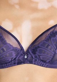 Violet nėrinio bralette su permatomis medžiagomis, gėlių raštais ir maža užtrauktuko detalė centre. Švelnus tekstūra ir reguliuojamos petnešėlės.