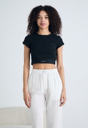 Black cropped t-shirt with short sleeves, featuring a Calvin Klein logo at the hem. Paired with white, pleated, loose-fitting trousers.