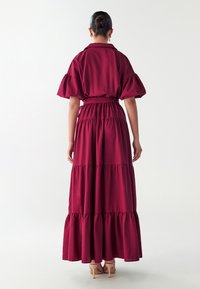 Dlouhé vínové šaty s krátkými puff rukávy, páskem v pase a vrstvenými volány na lemu. Hladká tkanina s jednobarevným vzhledem.