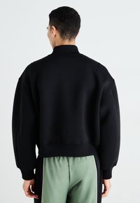 Sweat-shirt noir avec un col haut, coupe oversize et ourlet côtelé. Texture lisse, épaules tombantes et design minimaliste. Se termine au niveau de la taille.