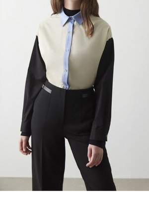 Žena v béžové košili s modrým knoflíkovým límcem a límečkem, černými dlouhými rukávy a černými kalhotami s vysokým pasem, stojící u bílé zdi.