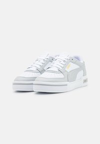 Een paar witte en lichtgrijze Puma-sneakers met witte veters, geperforeerde neus en een gouden Puma-logo aan de zijkant.