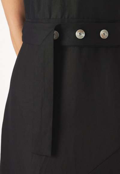 Détail d'une robe en textile noir avec une large ceinture nouée à la taille, fixée par trois boutons ronds de couleur claire. Texture lisse et mate.