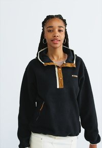 Μαύρο φλις πουλόβερ με καφέ κουμπί, κρεμ λεπτομέρειες και πλευρικό φερμουάρ. Διαθέτει κουκούλα και είναι φτιαγμένο από μαλακό, υφασμάτινο υλικό.