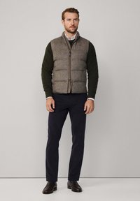 Padded prsluk od smešane tkanine s uzorkom houndstooth u smešnoj smešnoj boji, ispod kojeg je zeleni džemper s pletenim uzorkom. Uparen s tamnim hlačama i smešnim cipelama.