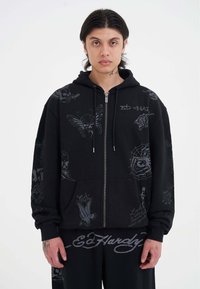 Ung mand iført sort Ed Hardy hættejakke og bukser med grå tatoveringsstil skelet, edderkop og tiger grafik, stående mod hvid baggrund.