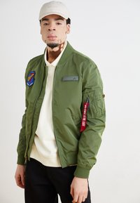 Olivzaļa bomber jaka ar rāvējslēdzēju, ribām piedurknēs un plāksteri uz krūtīm. Piedāvā sarkanu etiķeti uz piedurknes un gludu, spīdīgu tekstūru.