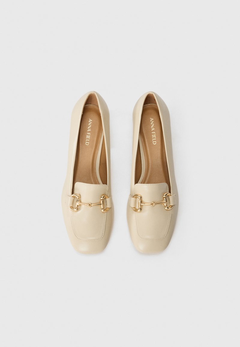 Mocassins en cuir beige avec des orteils carrés, présentant un accent en chaîne dorée sur le cou-de-pied et une finition lisse et texturée.