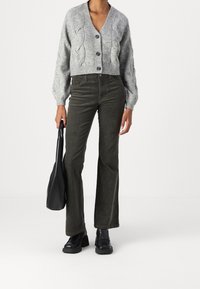 Šedý svetr s pleteným vzorem a velkými knoflíky, v kombinaci s tmavozelenými zvonovými kalhotami z kordu a černými platformovými botami, držící černou plátěnou tašku.