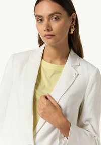 Weißer Blazer mit tailliertem Design und Reverskragen, über einem hellgelben T-Shirt getragen. Goldene auffällige Ohrringe fügen Details hinzu.