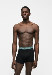 Μυώδης άνδρας στέκεται με χαλαρά χέρια, φοράει μαύρα μπόξερ Björn Borg με πράσινη ζώνη με λογότυπο, μπροστά σε απλό φωτεινό φόντο.