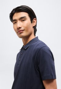 Νέος άνδρας με μαύρα μαλλιά που φοράει μια μπλε σκούρα υφασμάτινη κοντομάνικη πόλο μπλούζα, κοιτάζοντας ελαφρώς πλάγια με ουδέτερη έκφραση.