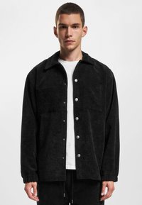 Black corduroy jacket with a button front, two chest pockets, and elastic cuffs. Styled over a white shirt, featuring a relaxed fit.