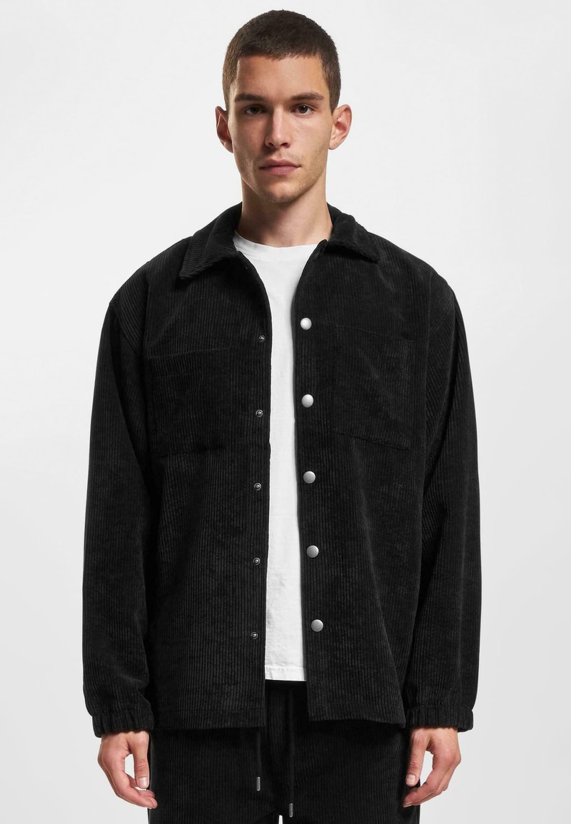 Veste en velours côtelé noir avec fermeture à boutons, deux poches poitrine et poignets élastiques. Portée par-dessus une chemise blanche, avec une coupe décontractée.