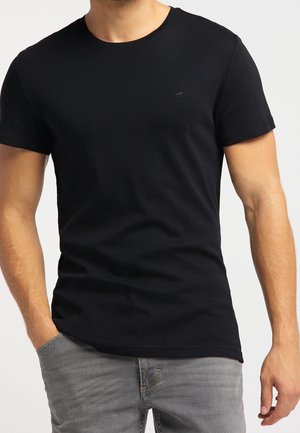 Maglietta nera in cotone con maniche corte, scollo a girocollo e logo discreto sul petto. Indossata con jeans grigi. Tessuto liscio.