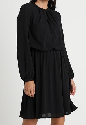 Vrouw die een zwarte jurk met lange mouwen tot op de knie draagt, met een geplooide halslijn en elastische manchetten, staand tegen een effen achtergrond.