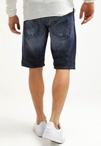 Man wearing dark blue denim shorts, white sneakers, and a light-colored long-sleeve shirt, shown from the back.