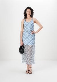 Blauw geruite maxi-jurk met een aansluitende lijfje en een uitlopende, doorschijnende zoom. Model houdt een zwarte tas vast en draagt zwarte sandalen met bandjes. Gladde stofstructuur.