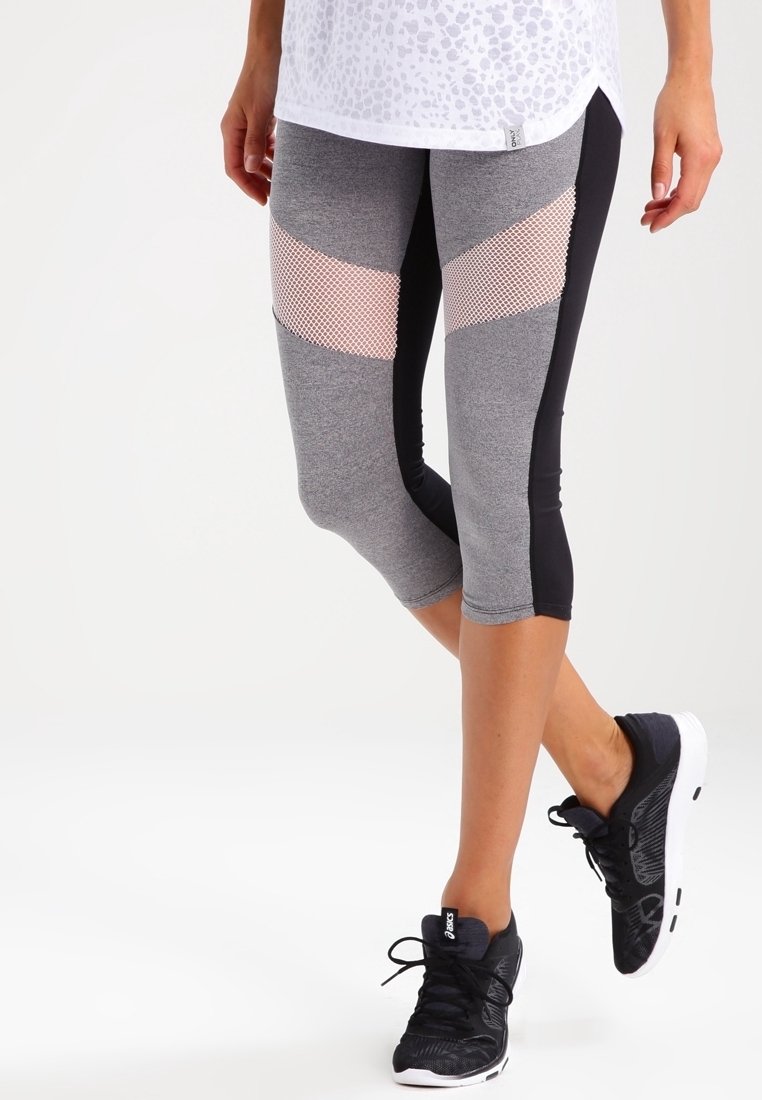 Jambes portant un legging court gris et noir avec des panneaux en mesh rose, assorties à des chaussures de course noires et blanches sur un fond uni.