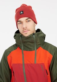 Kleurvolle jas in groen, rood en oranje, met een hoge kraag, trekkoorden en een logo patch. Draagt een rode gebreide beanie.