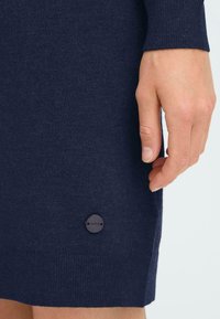 Marinblå stickad klänning med ribbade ärmar och nederkant, med en rund mörk knapp märkt "OXMO" på den nedre högra sidan. Slät textur.