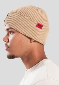 Profil boczny mężczyzny w beżowej dzianinowej czapce z czerwonym napisem "WRONG FRIENDS" oraz białej koszulce z okrągłym dekoltem.