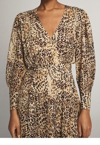 Długo rękawowa sukienka w kolorze beżowym i brązowym, w zwierzęcy wzór. Posiada głęboki dekolt w kształcie litery V, wcięcie w talii oraz miękką, lejącą się strukturę materiału.