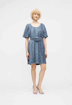 Γυναίκα που στέκεται φορώντας ένα μπλε φόρεμα με κοντά μανίκια και υφή, με ζώνη που έχει υφασμάτινο λουλούδι, σε συνδυασμό με ανοιχτό γκρι ψηλοτάκουνα σανδάλια.