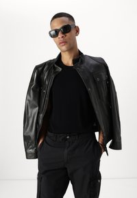Veste en cuir noire avec des boutons-pression, portée sur un shirt en maille noire, associée à un pantalon cargo noir. Les accessoires comprennent des lunettes de soleil sombres.